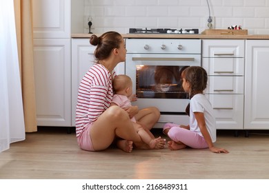 Horizontal shot of happy woman with bun hairstyle wearing striped shirt sitting on floor in kitchen with her kids, mother and her kids waiting for tasty pie, baking in oven. - Powered by Shutterstock