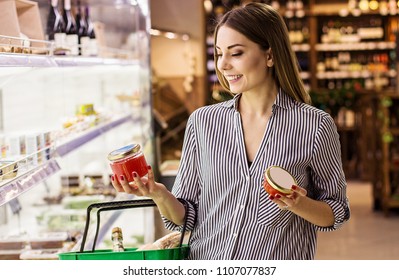 Horizontal Shot Happy Woman Reading Label With Price At Caviar Jar While Standing At Seafood Supermarket. Beautiful Young Girl Choosing Red Caviar While Doing Shopping At Grocery Shop. Fish Appetizer