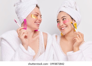 Horizontal shot of happy women look at each other smile toothily enjoy beauty procedures at home hold cosmetic brush apply moisturising patches under eyes have healthy well cared skin wear bathrobes - Powered by Shutterstock