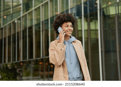 Horizontal Shot Of Happy Young Woman Makes Telephone Call In Roaming Wears Casual Sweatshirt And Beige Jacket Had Glad Expression Walks Against Blurred City Building Enjoys Smartphone Talking