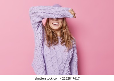 Horizontal Shot Of Happy Young Woman Covers Eyes With Arm Wears Loose Knitted Sweater Tries To Hide Face Stays Positive Isolated Over Pink Background. Cheerful Female Model Awaits For Surprise