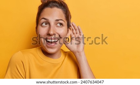 Horizontal shot of healthy young cheerful woman keeps hand near ear tries to overhear rumors listens private information with interest has curious expression isolated over yellow background.