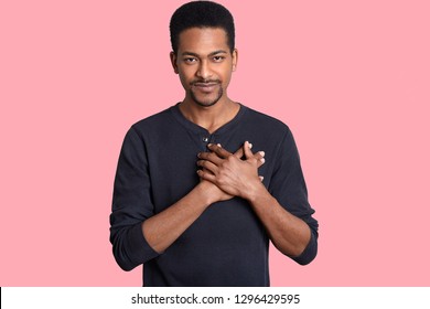 Horizontal shot of impressed touched man holds hands on chest, expresses thankfullness, hears heart piercing words, dressed in casual jumper, isolated over pink background. People and gratitude