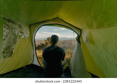 Horizontal shot from inside the green tent of a black male hiker getting up at sunrise during camping trip with view of the snowy mountaintop and lush greenery