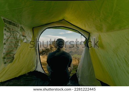 Horizontal shot from inside the green tent of a black male hiker getting up at sunrise during camping trip with view of the snowy mountaintop and lush greenery