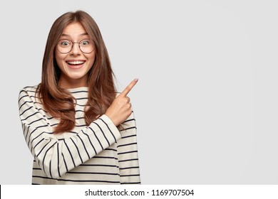 Horizontal shot of joyful pleased good looking youngster points at upper right corner with index finger, invites you go there, dressed in casual clothes, poses against white background, blank space - Shutterstock ID 1169707504
