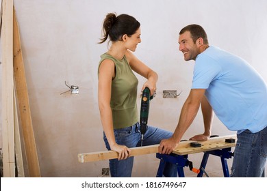 Horizontal Shot Of A Joyous Couple Doing DIY At Home With Woman Drilling Hole With Power Drill On Workbench And Man Assisting.