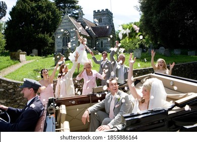 Horizontal Shot Of A Jubilant Wedding Party Throwing Confetti On Bride And Groom In A Convertible Vintage Car By The Church On A Sunny Day.