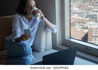 Horizontal Shot Latina Woman Working From Home, Drinking Coffee, While Looking At Smart Phone, With Copy Space.