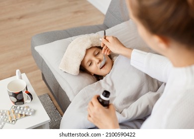 Horizontal Shot Of Little Sick Girl Lying On Sofa, Mother Giving Her Spoon With Syrup, Family Posing In Light Room, Kid Taking Medicine From Coughing Or High Temperature.