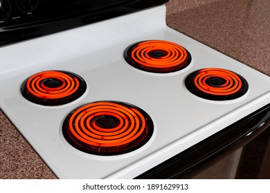 Horizontal Shot Looking Down On A White Stove Top With Four Burners Glowing Orange.