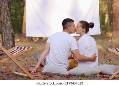 Horizontal Shot Of Loving Romantic Couple Sitting In The Forest With Overhead Projector With Empty Screen For Promotional Text, Wife And Husband Hugging And Kissing, Celebrating Their Anniversary.