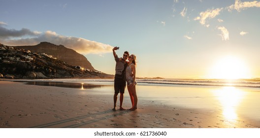 Horizontal shot of loving young couple taking self portrait at the beach. Man and woman on sea shore taking selfie with mobile phone. - Powered by Shutterstock