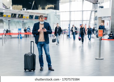 Horizontal Shot Of Man Poses In Crowded Airport, Uses Cell Phone, Checks Time Of Flight Online, Stands Near Suitcase, Wears Medical Mask During Coronavirus Crisis. Health Care, Traveling Concept