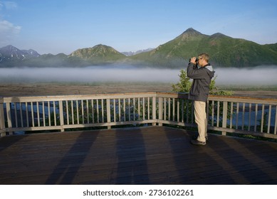 the horizontal shot of a man using binoculars to look at the foggy mountains beyond the fence on a sunny day