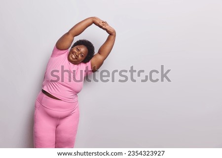 Horizontal shot of overweight woman has regular training to loose weight keeps arms raised up and leans on right dressed in pink sportswear isolated over white background copy space for your text