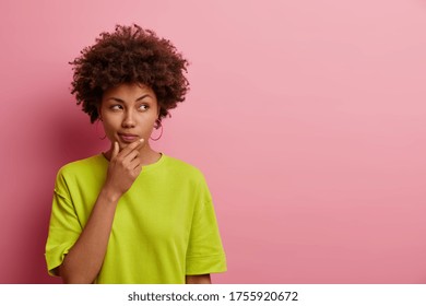 Horizontal Shot Of Pleasant Looking Afro American Woman Holds Chin And Looks Pensively Away, Thinks About Great Plan, Has New Ideas In Mind, Wears Bright Green T Shirt, Poses Over Rosy Wall Copy Space