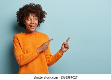 Horizontal shot pleased dark skinned female with Afro haircut, points away with both fore fingers, shows blank space for your promotion, isolated over blue background. People, advertisement concept