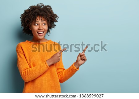 Horizontal shot pleased dark skinned female with Afro haircut, points away with both fore fingers, shows blank space for your promotion, isolated over blue background. People, advertisement concept