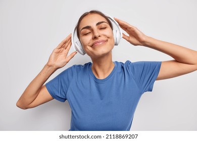 Horizontal shot of pleased dreamy young woman wears stereo headphones on ears enjoys listening music keeps eyes closed wears casual blue t shirt isolated over white background. People and hobby