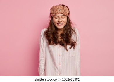 Horizontal Shot Of Pleased Young European Woman Closes Eyes And Smiles Gently, Wakes Up In Good Mood After Seeing Pleasant Dreams At Night, Dressed In Casual Domestic Clothes, Funny Eyemask On Head