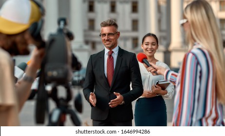 Horizontal Shot Of Politician Giving Interview To Journalists. People Making Interview Using Digital Camera And Equipment Set At Outdoor Location. Selective Focus. Front View