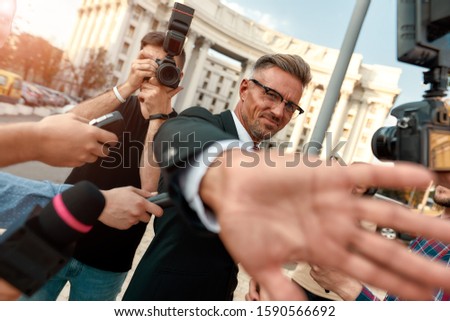 Horizontal shot of politician trying to cover camera lens with his hand while refusing to give interview. Reporters want to interview him using microphones and equipment. Selective focus. Dutch angle