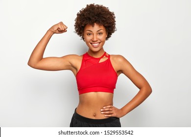 Horizontal shot of positive dark skinned woman shows biceps, demonstrates strong hand, has slim figure, wears sport bra, smiles pleasantly, isolated over white background. People and strength