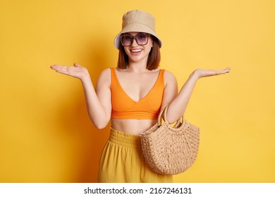 Horizontal shot of positive young young woman wearing swimsuit, shorts and panama, posing isolated over yellow background, standing spread arms, being not sure, don't know.