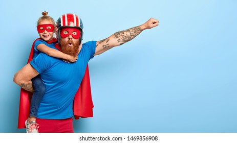 Horizontal Shot Of Powerful Man Makes Fly Gesture, Gives Piggyback To Small Daughter Dressed In Superhero Suit, Have Supernatural Abilities, Enjoy Free Time Together. Real Heroes Defend You.