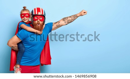 Horizontal shot of powerful man makes fly gesture, gives piggyback to small daughter dressed in superhero suit, have supernatural abilities, enjoy free time together. Real heroes defend you.