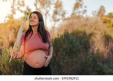 Horizontal Shot Of A Pregnant Latina Woman In The Middle Of Nature, Smelling A Feather Reed Herb While Smiling And Holding Her Belly. Copy Space