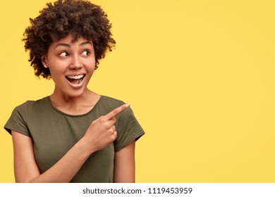 Horizontal shot of prettty young joyful African American female points with fore finger away, indicates copy space on blank wall, isolated over yellow background. Check this out. Ethnicity concept