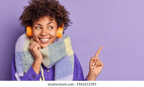 Horizontal Shot Of Pretty Afro American Woman With Broad Smile White Teeth Presents Something Points At Upper Right Corner Listens Music Via Headphones Wears Warm Scarf Around Neck. Look There