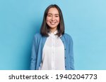 Horizontal shot of pretty Asian woman with dark hair smiles pleasantly looks directly at camera has toothy smile wears white shirt and jumper isolated over blue background. Emotions concept.