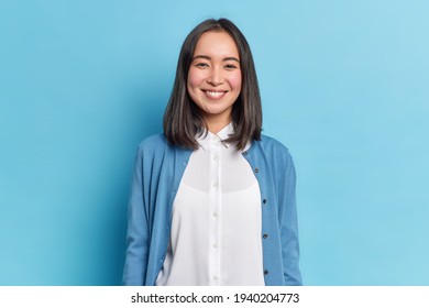 Horizontal shot of pretty Asian woman with dark hair smiles pleasantly looks directly at camera has toothy smile wears white shirt and jumper isolated over blue background. Emotions concept. - Shutterstock ID 1940204773