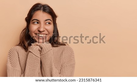 Horizontal shot of pretty dark haired woman looks aside has toothy smile keeps hands under chin dressed in loose knitted jumper poses against brown background copy space for your promotional content