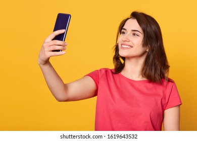 Horizontal shot of pretty girl taking self portrait with her smart phone, adorable lady looking devie's at camera with charming smile, attractive model posing isolated over yellow studio background.