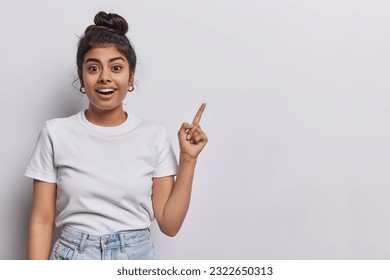 Horizontal shot of pretty surprised cheerful young woman pointing to empty copy space advertises product or tells about awesome offer dressed in casual clothing isolated over white background