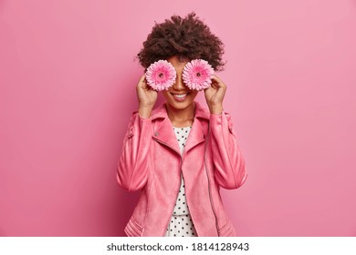 Horizontal Shot Of Pretty Woman Has Afro Hair Holds Two Gerbera Daisy In Front Of Eyes And Smiles Broadly, Has Fun. Fashionable Lady Enjoys Spring Time, Pleasant Fragrance. Florist With Flowers