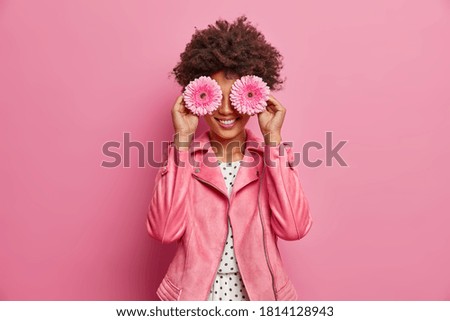 Horizontal shot of pretty woman has Afro hair holds two gerbera daisy in front of eyes and smiles broadly, has fun. Fashionable lady enjoys spring time, pleasant fragrance. Florist with flowers