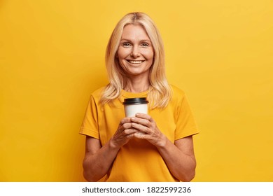 Horizontal Shot Of Pretty Wrinkled Middle Aged Woman Holds Disposable Cup Of Coffee Enjoys Free Time And Looks Happily At Camera Dressed In Casual T Shirt Isolated On Vivid Yellow Background