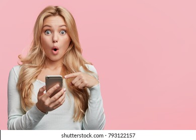 Horizontal shot of pretty young female with long hair looks with bugged eyes, holds modern smart phone, recieves unexpcted message from friend, reads reminder, isolated over pink background.