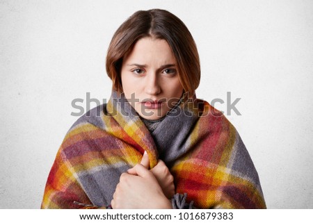 Horizontal shot of pretty young woman feels cold, wrapped in wool coverlet, tries to warm herself, isolated over white background. Adorable female trembles after walk in frosty weather outdoor
