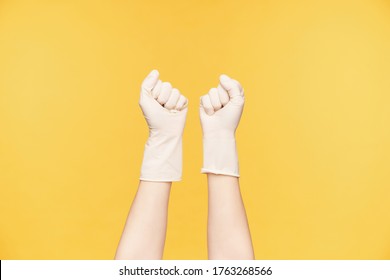 Horizontal shot of raised young woman's hands in rubber gloves clenching fists while posing over orange background. Cleaning and house care concept
