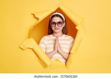 Horizontal Shot Of Relaxed Woman Wearing Striped Shirt And Hair Band Posing In Yellow Paper Hole, Practicing Yoga, Keeps Palms Together In Praying Gesture.