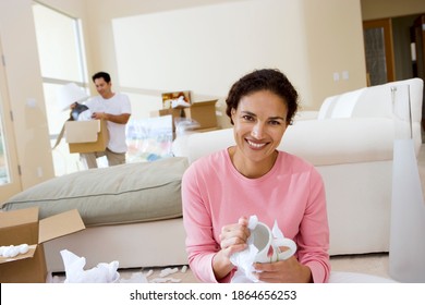 Horizontal Shot With Selective Focus On The Woman In Foreground Unwrapping Glassware Smiling At The Camera With Her Husband In Background Unboxing A Lamp.