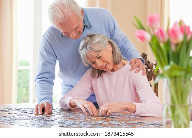 Horizontal shot of a senior couple assembling a jigsaw puzzle at a table indoors with the man's hand on wife's shoulder and a flower vase in the foreground. - Powered by Shutterstock