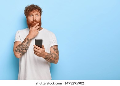 Horizontal shot of serious contemplative adult man touches thick red beard, holds mobile phone, browses newsfeed online, thinks over recent news, has tattooed arms, wears casual white t shirt - Shutterstock ID 1493295902