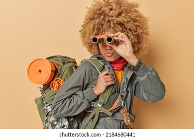 Horizontal shot of serious curly woman camper explores unknown place focused into distance keeps binoculars near eyes carries heavy rucksack travels in mountains poses against beige background